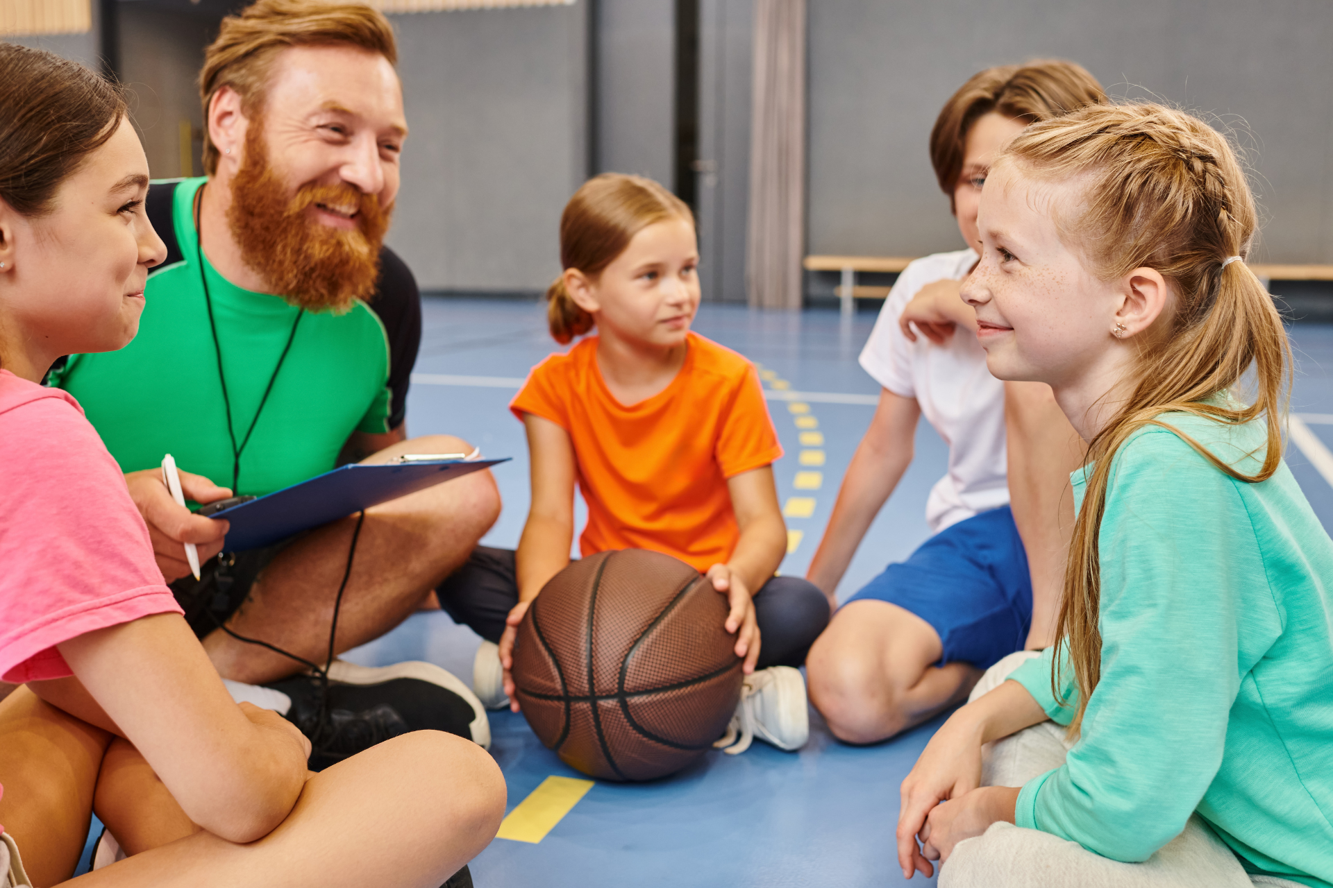 Students sitting on a gym floor with a teacher and a basketball