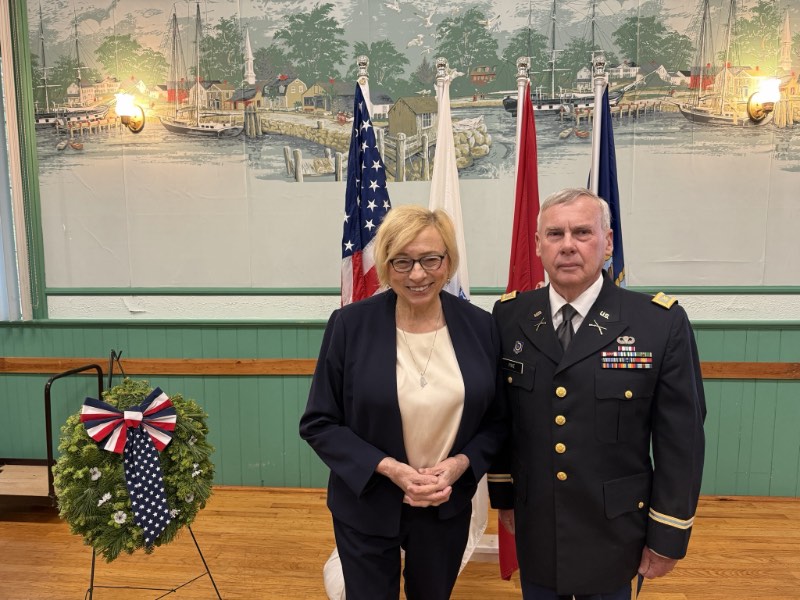 Governor Mills and keynote speaker Major Robert Pike standing in front of the US and State of Maine flags, and a wreath with a red, white, and blue ribbon